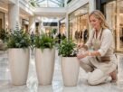 Mulher sorridente usando avental bege agachada cuidando de plantas em um kit com três vasos de polietileno grandes e cilíndricos em tom neutro, posicionados em um corredor de shopping com piso de mármore.