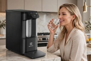 Mulher loira sorridente na cozinha, vestindo blusa bege, bebendo um copo de água fresca ao lado de um purificador de água Electrolux preto moderno instalado sobre uma bancada de mármore.