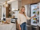 Uma mulher loira e sorridente, vestindo um suéter branco e jeans, está em pé em uma cozinha moderna e luxuosa com armários de madeira e bancadas de mármore. Ela está com as mãos na alça de um refrigerador duplex Electrolux IF41 Frost Free de cor branca com portas fechadas, que está embutido nos armários. Uma ilha de mármore com pães e limões está em primeiro
